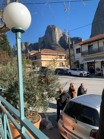 This image captures a bright, sunny street scene in Kastraki, Greece, with dramatic rock formations characteristic of Meteora dominating the background under a clear blue sky. The photo appears to have been taken from a slightly elevated position, possibly a balcony, as indicated by a turquoise railing in the lower right foreground.
In the immediate foreground on the left, a large, spherical white street lamp is prominent on a turquoise pole. Below, the street and sidewalk are bustling with activity. A group of approximately three to four adults is gathered; one man in a dark jacket is seen pointing towards something out of frame or in the distance. Another man in a dark jacket is bending down next to a silver Toyota Yaris car, seemingly attending to a small, fluffy white dog wearing a light-colored sweater, which is on a leash near a planter. A woman in a brown jacket stands nearby.
Several cars are parked along the street, including the silver Toyota Yaris, which has "Yaris" written on its rear and a Greek license plate starting with "ITY". A white car and a darker vehicle are also visible further down the street. The buildings lining the street feature traditional architecture with balconies, tiled roofs, and storefronts. One sign on a building appears to say "Bakery," though it is partially obscured. Utility poles and overhead power lines are also visible against the sky. The overall impression is a bustling small town or village scene, with the towering natural landscape creating a striking contrast.