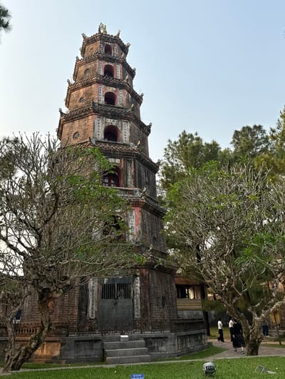 The image captures the iconic Thien Mu Pagoda, located in Kim Long Ward, Thua Thien Hue, Vietnam. The pagoda, constructed with brown brick, rises prominently against a pale sky. It is composed of tiered segments, each detailed with architectural ornaments and inscriptions, and a small arched window in each tier. A bare tree with sparse foliage partially obscures the pagoda's base, framing the structure with its branches. To the right, a glimpse of a grassy area and a pathway occupied by several people can be observed. The scene is illuminated by soft, natural light, emphasizing the ancient architecture.