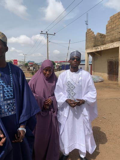 The image shows three people outside in Naraguta Hausawa, Nigeria. A man in a white, intricately embroidered traditional garment and sunglasses is holding a mobile phone. Next to him is a woman in a light purple hijab and dress, holding a small cup or glass. Partially visible at the bottom is a man in a dark blue robe with blue embroidery. The background includes a partially constructed brick building, utility poles with wires, a street, and buildings in the distance. The ground appears to be dirt or gravel. The sky is light blue with some clouds.