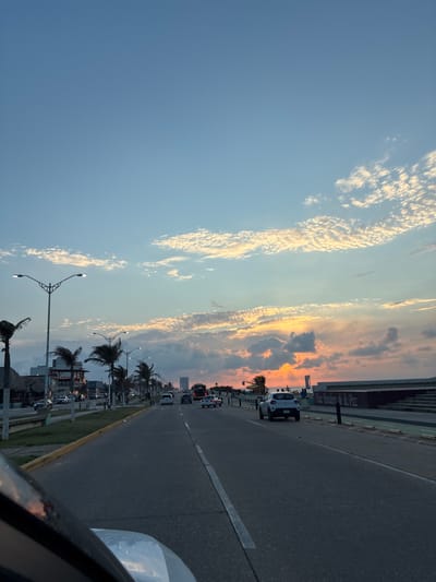 The image depicts a view from a vehicle driving along a road in Coatzacoalcos, Mexico. The road appears to be a multi-lane highway with a median or barrier. Palm trees line the side of the road. Buildings and other structures are visible in the background, including what may be a restaurant or commercial establishment. Traffic is present on the road, with several cars visible. The sky above features scattered clouds and a gradient of light, suggesting either sunrise or sunset. A sign saying "Villas Baneras de la Paz" can be seen along the road.