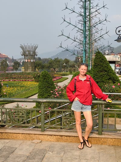 A female individual stands posing in the Sa Pa Lake Park and Flower Gardens in Sa Pa, Vietnam. She is positioned behind a dark green metal railing with ornamental circular motifs, resting her right arm on it while holding a yellow smartphone. Her left hand is in her pocket. She wears a red full-zip jacket with a visible "The North Face" logo, a grey t-shirt, light blue denim shorts, and black thong-style sandals. Sunglasses are pushed onto the top of her head.
Behind the individual and the railing, a meticulously maintained public garden unfolds, featuring paved walkways, conical evergreen shrubs, and flower beds densely planted with yellow flowers and pink roses. In the mid-ground, a tall, stylized metal tree structure, painted green and blue with small decorative elements, stands to the right. A prominent, large metal sculpture resembling a crown or stylized lotus, adorned with patterned panels at its base, is situated further back in the garden, adjacent to Sa Pa Lake (Hồ Sa Pa). Traditional-style buildings with multi-tiered red roofs are visible on the far side of the lake, against a backdrop of hazy, distant mountains under a light grey sky. Vehicles are partially visible on a road in the far right background. The foreground consists of grey stone paving and a textured golden-brown curb.