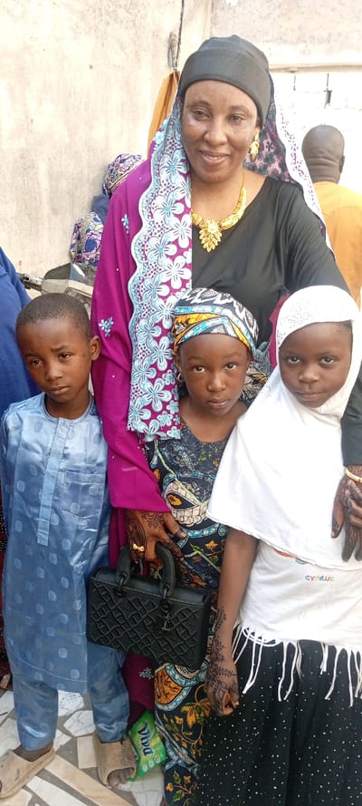 A group of four individuals, consisting of one adult woman and three children, are standing outdoors against a light-colored wall in Dan Maliki, Nigeria. The adult woman, positioned centrally, wears a black top, a magenta and light blue patterned head covering, a gold-colored necklace, and earrings, with henna designs visible on her hands. She smiles and holds a black, quilted handbag in her right hand, while her left arm is around the shoulder of a young girl to her right. To the adult woman's left stands a young boy wearing a light blue patterned garment and light brown sandals, looking directly at the camera with a neutral expression. Between the adult woman and the boy stands a young girl in a dark patterned dress and a blue, yellow, and black patterned head covering, also looking at the camera with a neutral expression. Her hands display henna, and she wears small hoop earrings. To the adult woman's right is another young girl, wearing a white head covering, a white top with "CYPRUS" lettering, and a black polka-dotted skirt with white fringes. Her hands also show henna, and she looks directly at the camera with a neutral expression. A green "Viva" package lies on the tiled ground near the boy's feet. Partially visible figures include a person in a blue garment behind the boy and the back of an individual in an orange garment behind the adult woman on the right.