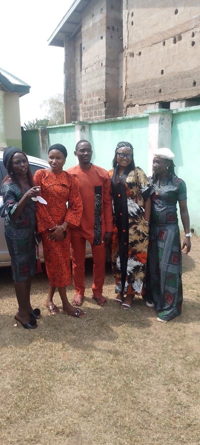 The image displays five individuals—four women and one man—posing outdoors in daylight against a backdrop in Ohafia, Nigeria. They stand on a patch of dry, light-brown grass.
All individuals are adults and are dressed in brightly patterned attire, characteristic of traditional or celebratory African prints. From left to right: a woman in a dark patterned dress with a black headwrap and high-heeled sandals, holding a white tissue; a woman in an orange and black patterned top and skirt with a dark headwrap and brown flat sandals; a man in an orange long-sleeved tunic with a black patterned stripe, matching trousers, and brown open-toed sandals; a woman in a long, flowing multi-colored kaftan (black, orange, white patterns) wearing dark sunglasses, a dark headwrap, and white open-toed sandals; and finally, a woman in a patterned short-sleeved top and matching long skirt, wearing a white cap and dark shoes or sneakers.
In the background, a light-colored car is partially visible behind the leftmost woman. A low, light-green wall with concrete pillars runs behind the group, separating them from a large, multi-story building. This background building appears to be constructed of weathered concrete blocks, showing exposed blockwork and several small holes, topped with a dark roof overhang. A portion of another building with a green roof is visible on the far left. The bright lighting suggests a clear, sunny day. The individuals mostly have pleasant or neutral expressions, indicating a relaxed atmosphere. No visible text is present in the image.