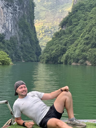 A landscape scene in Đồng Văn Commune, Vietnam, shows a Caucasian male lying on his side along a lake's edge. The man wears a light gray t-shirt, black shorts, a gray baseball cap, and gray running shoes with gray socks. His arms are positioned with his fists clasped together. He wears a watch on his left wrist and a woven bracelet on his right. He is positioned near a white painted metal railing.
To the left of the lake is a verdant mountain covered in lush vegetation, with areas of exposed rock. The lake's water appears green and reflects the surrounding landscape. The overall scene suggests a natural, outdoor setting.