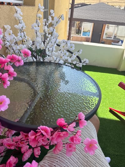 The image shows an outdoor setting with artificial flowers, a glass-top table, and a person's leg. The scene appears to be a balcony or patio, suggested by the railing and the building in the background. The artificial flowers, in both white and pink, are arranged around the table. The table has a dark metal frame and a textured glass top, reflecting the surroundings. A person's leg, clad in light-colored textured fabric (possibly a blanket or clothing), is visible on the right side of the frame, resting on a surface that looks like artificial grass. In the background, parts of a building and windows are visible, suggesting an urban or semi-urban environment. The lighting suggests it is daytime, likely with sunlight casting shadows, particularly from red objects not fully in frame. The overall mood is relaxed and summery. No visible text is present in the image.