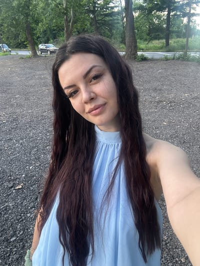 A young woman with long, dark, wavy hair, wearing a light blue halter top, is taking a selfie outdoors. She is standing in an area covered with dark gravel. Behind her, a road is visible with trees and foliage lining the sides. Two cars are parked on the side of the road in the background. The lighting suggests it is daytime, possibly late afternoon due to the soft quality of the light. The overall setting appears to be a rural or semi-rural area on the outskirts of a city. The location is identified as Zlati Voyvoda, Bulgaria.