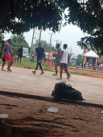 In Ukukwa, Nigeria, a group of young men are engaged in a lively game of basketball on an outdoor court. The scene is set beneath the shade of lush green trees, with a cloudy sky overhead suggesting a potentially overcast day. In the foreground, a pile of dark backpacks rests on the reddish-brown dirt bordering the court.
The main subjects are four young men actively playing basketball. One individual in a blue and white jersey with red shorts is dribbling the ball, while another in a purple and yellow jersey, holding the ball, appears to be passing or shooting. Two other players, one in a black t-shirt and black leggings, and another in a white t-shirt and dark shorts, are actively defending or moving on the court.
In the background, several other individuals are gathered, observing the game or socializing. A small, light-colored building with a distinctive white roof stands to the right, possibly a structure on the sports facility grounds. The presence of utility poles and overhead wires indicates an urban or semi-urban setting. A white board with visible text is placed near the basketball hoop on the left side of the court. The overall atmosphere appears casual and energetic, typical of a friendly pickup basketball game.