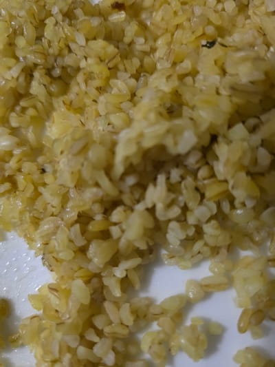 The image shows a close-up, overhead view of cooked bulgur wheat. The grains are light brown and appear slightly oily. Some individual grains are visible, as well as clumps where they have stuck together. There are also some darker specks scattered throughout the bulgur, which may be spices or small pieces of debris. The bulgur is served on a white plate, with some of the grains spilling over the edge. The lighting is somewhat dim, and the focus is sharp on the bulgur. This image is not geo-tagged and cannot be placed in Başakşehir, Turkey.