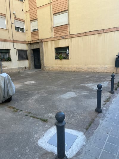 The image depicts an outdoor courtyard in the city of Castelló De La Plana, Spain, with a building facade on the left and rear. The building is constructed with beige stucco and features horizontal banding in a darker shade of beige. Several windows with white blinds are visible, some with green metal security grates. A dark metal door is situated in the center of the building's ground floor.
The courtyard floor is paved with concrete, showing some wear and discoloration. A motorcycle covered with a silver tarp is parked on the left side of the courtyard. Several black bollards with spherical tops are positioned in the courtyard, one closer to the foreground and two further back. A trash receptacle is visible on the far right edge of the image, near the sidewalk. The sidewalk itself is paved with square grey tiles. A small black and white flag with a graphic is visible in a window of the building on the right. The scene appears to be an ordinary residential or communal outdoor space.