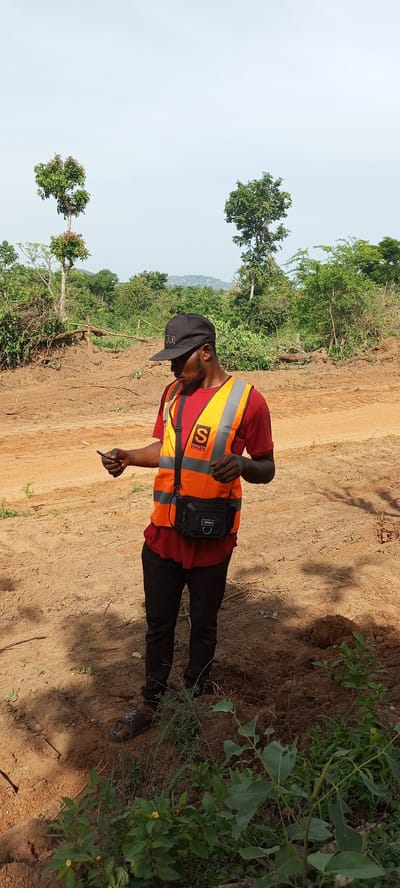 A man stands outdoors in a cleared, earthy area, likely in Otunade, Nigeria. He is of Black ethnicity, wearing a black baseball cap with "OLD" visible on the front, a red short-sleeved t-shirt, and an orange high-visibility vest with reflective stripes. On the left side of his vest, a logo with a large "S" and "SWALBYS" underneath is visible. He also wears black pants and black open-toed sandals. A black waist bag with "SPORT" written on it is worn across his chest. He is holding a small dark object, likely a smartphone, in his left hand and is looking down at it with a focused expression.

The setting appears to be a construction site or recently cleared land, characterized by reddish-brown, uneven dirt with visible tracks or disturbances. Patches of green vegetation and small plants are scattered on the ground, particularly in the foreground. In the mid-ground and background, numerous trees and dense foliage are present, with distant hills visible under a pale blue, slightly hazy sky. The lighting suggests it is daytime, possibly mid-morning or early afternoon, with shadows cast by the man indicating the sun's position.