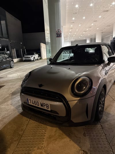 The scene depicts a silver Mini Cooper convertible, license plate '7460 MPB', parked at night on a light-colored concrete surface with raised tactile paving elements, within the Polígon Industrial De Son Castello, Spain. The vehicle is angled slightly towards the left, with its front fascia, left headlight, and a portion of the hood prominently featured. The car's soft top appears lowered, and the interior is dark. To the right of the Mini, a large glass window reflects numerous interior overhead lights from a brightly lit commercial space, likely a showroom or dealership. A red and white "Prohibido el Paso" (No Entry) sign is affixed to a vertical glass column or pillar separating the glass panels. In the background, to the left, a dark grey vehicle is partially visible, parked on a similar concrete surface. Further back, a white Mini Cooper convertible is parked in front of a dark, utilitarian building facade under the night sky. No individuals are visible in the image. The lighting is primarily artificial, originating from the building interior and possibly streetlights off-frame, casting shadows on the ground.