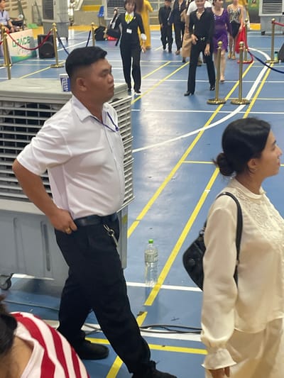 In Thuận An, Vietnam, a man in a white short-sleeved collared shirt and black pants stands to the left of the frame, looking to his right. He has dark hair and is wearing a gold ring on his right hand. A set of keys with a silver fob is attached to his belt. To his right, a woman with dark hair pulled back in a bun is visible from the shoulders up, wearing a cream-colored blouse with ruffled cuffs and embroidered detailing on the neckline. She has a black backpack slung over her right shoulder. In the background, other individuals, some in formal wear, are present on a blue floor marked with yellow lines. To the far left, a large gray industrial fan is visible. A plastic water bottle with a green cap rests on the floor in the middle ground. The scene appears to be an indoor event or competition.