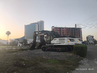 A large white excavator with "HIDROMEK" branding sits on a gravelly patch of land. In the background are several buildings, including a tall, modern hotel and a partially constructed brick building. Overhead electrical wires are partially visible. The sky is pale and suggests early morning light. The date "23/11/2025 06:41" is stamped in the lower right corner of the image.