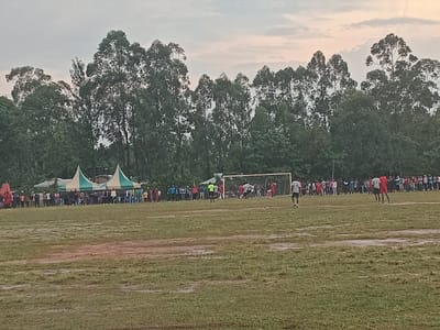A wide shot shows a soccer game in progress on a muddy field, with spectators lining the sidelines. Tall trees form the backdrop, with hints of tents or structures visible behind the crowd. The sky is overcast, suggesting possible rain or a recent downpour. Players are positioned around the goal area, and the focus is on the action taking place near the net.