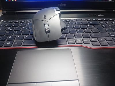 The image shows a close-up, high-angle view of a black laptop with a gray mouse placed on the keyboard. The laptop screen is visible at the top, displaying a portion of a graphical user interface and the word "FUJITSU." The keyboard is a standard QWERTZ layout, with keys such as "Esc," "1," "2," "3," "4," "Q," "W," "E," "R," "A," "S," "D," "F," "Z," "X," "C," and various function keys and modifiers visible. The mouse is ergonomic and gray, with a prominent scroll wheel and several buttons. The red trim around the keyboard area is also visible. The laptop's trackpad is partially visible in the foreground, to the left of the keyboard. The scene appears to be indoors, with artificial lighting. There are no people visible in the image. The location of Bohodukhiv, Ukraine is not indicated by any visual cues in the image.