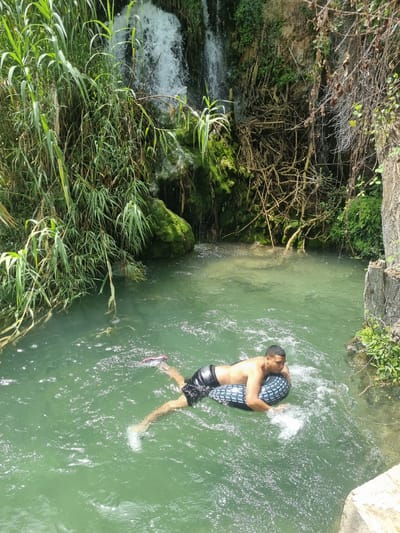 A young man is swimming in a shallow river with a tire inner tube. He is holding the tube and is in a playful mood. The water is clear and green, and the surrounding environment is lush with vegetation. The sun is shining brightly, indicating it is a warm, sunny day. The location appears to be a natural setting, possibly a park or a nature reserve. The man is wearing swim trunks and a pair of sneakers. The scene is peaceful and serene, suggesting a relaxing day out in nature. There are no visible text in the image.