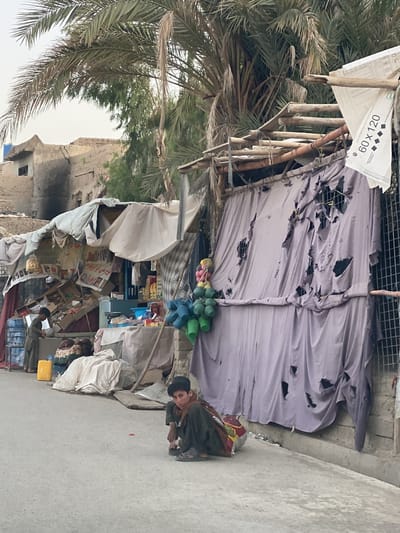 This street-level photograph depicts a bustling, yet rudimentary, scene in Arghandab, Afghanistan, under a light, hazy sky. The setting is characterized by makeshift stalls, worn buildings, and prominent palm trees.

In the immediate foreground, a young boy is crouched on the concrete pavement, looking directly towards the viewer. He wears dark, traditional clothing and sandals, with a colorful, patterned bag or cloth resting beside him.

Behind him, to the left, another boy stands near a makeshift stall constructed from wooden poles and draped fabric awnings. This stall displays various goods, including stacked boxes and packaged items, suggesting a small shop or market stand. A bright yellow jerrycan is visible on the ground near this stall. Beyond this, dilapidated buildings, possibly made of mud brick, show signs of damage or age, with one exhibiting a large, dark, possibly burnt area.

To the right, another structure features a wooden frame and a large, faded purple-grey fabric, heavily torn and holed, serving as a curtain or wall. Colorful plastic items, possibly toys or small containers, hang from a string in front of this fabric. Next to this structure, a metal grid fence is visible. A white piece of material attached to the right structure displays the black text "60 x 120" along with several diamond shapes.

Large palm trees with abundant fronds dominate the upper part of the image, extending across the sky and providing shade. The overall atmosphere suggests daily life in a resilient community, likely in an area facing economic challenges. The diffused light indicates either early morning or late afternoon.