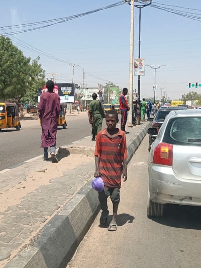 The scene depicts a sunny daytime street in Hotoro South, Nigeria. In the immediate foreground, a young male, appearing to be of African descent, walks towards the camera on a dirt patch alongside a concrete curb. He wears a faded red and black striped t-shirt, dark trousers, and sandals, holding a purple plastic bowl in his right hand. His gaze is directed forward. To his right, a silver hatchback car (possibly a Toyota Corolla) is parked parallel to the curb, its rear section visible.
Further back on the left, another male, also appearing African, walks away from the camera on a paved sidewalk, wearing a long, loose dark red garment over blue jeans. Adjacent to the road on the left, a yellow auto-rickshaw is partially visible. Centered in the midground, a man in an olive green uniform walks away from the camera, seemingly carrying coiled ropes. To his right, another male in a red and blue tracksuit stands near a light pole, facing generally towards the right. Several other pedestrians are visible further down the street on the right side of the road.
The environment features a paved road separated from a paved sidewalk by a concrete curb; the sidewalk exhibits patches of bare dirt and sand. Utility poles with overhead power lines crisscross the clear blue sky. A large digital billboard in the midground on the left displays "BABA / BOS GO AGAIN 2027" with political imagery. Other various billboards and signs are present along the road, some with green motifs. Green foliage from trees is visible on the left and in the distant background. In the far distance on the right, traffic lights are visible.