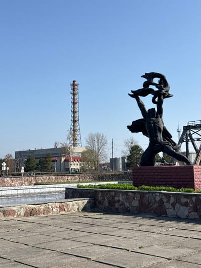 The image depicts a monument, most likely the "Those Who Saved the World" monument, in Pryp'yat', Ukraine, with the Chernobyl Nuclear Power Plant visible in the background. The sculpture is bronze and features a man reaching towards the sky, possibly holding a child or symbol of life. The statue stands on a stone and brick pedestal. A tall, rusty metal structure, part of the nuclear plant, rises prominently behind the monument. The sky is clear blue. The foreground shows a paved walkway, suggesting a public space. The scene is still and silent, conveying a somber atmosphere.