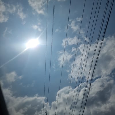 The image captures a bright, sunny sky with scattered white and gray clouds, viewed from below. A prominent sunburst effect emanates from the upper left quadrant of the image, indicating a strong light source. A series of dark, parallel wires, likely power or telephone lines, stretch diagonally across the frame from the bottom left to the upper right. These wires are held in place by visible attachments, suggesting they are part of an overhead utility infrastructure. The ground is not visible, and the perspective is upward-looking. The scene is set in Anheung-myeon, South Korea.