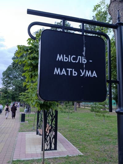 The image shows a black square sign with white Cyrillic text, "МЫСЛЬ - МАТЬ УМА" (Thought - Mother of the Mind), mounted on a black metal pole in a park. The sign has some white smudges on its surface. Behind the sign, a paved walkway with people walking can be seen, lined with green trees and grass. The overall setting appears to be an outdoor public space, possibly a park in the city of Kelermesskaya, Russia, given the presence of Cyrillic text and the typical urban park environment. The sky is overcast.