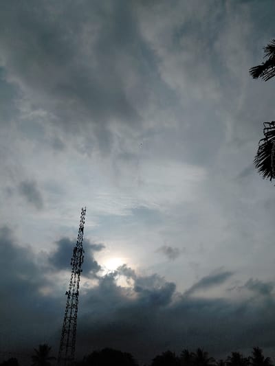 The scene is located in Mns Alue, Indonesia, under an overcast sky. A tall, lattice-structure communication tower stands on the left side of the frame, silhouetted against the sky. The sun is partially visible and diffused by dense, dark gray clouds positioned behind and around it, creating a bright central glow. The sky is predominantly filled with varying shades of gray clouds, some appearing heavy and dark. The lower portion of the image contains dark silhouettes of vegetation, including the tops of trees and distinct palm fronds visible on the far right. No people or specific activities are present in the image.