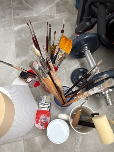 The image depicts an overhead view of art supplies and workout equipment arranged on a tiled floor. In the foreground, a collection of paintbrushes in various sizes and states of use are held in a blue cylindrical container. Some brushes are clean, while others show traces of paint. Adjacent to the container, a white roll of textured material, possibly canvas or paper, is partially visible on the left. To the right of the brushes, a white mug contains several sculpting tools and a small foam roller. A white lid for a container is also present near the mug. In the background, weightlifting equipment, including dumbbells and part of a weight machine, are visible. A small bottle with a black cap and amber liquid, possibly paint or varnish, is positioned among the paintbrushes. The scene appears to be set in a home studio or workshop environment in Mesogi, Cyprus. There are no people or active interactions visible in the image. The lighting suggests an indoor setting, possibly with natural light from above. The floor is made of grey tiles with subtle veining.
