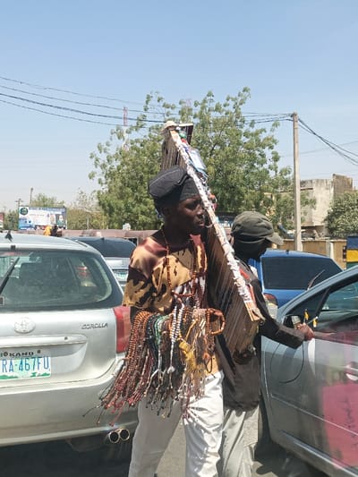 The image depicts a street scene in Hotoro South, Nigeria, under clear daylight. A dark-skinned male, centrally positioned, is seen navigating between vehicles. He wears a brown and tan patterned short-sleeved shirt, white trousers, and a black cap. Numerous strands of various colored beads, appearing to be merchandise, are draped over his left shoulder and chest. He carries a large, light-colored wooden display board vertically against his right shoulder, featuring multiple small items, possibly pictures or trinkets, affixed to its surface. To his right, another dark-skinned male, wearing a dark shirt and a dark cap, stands beside a silver car, with a partial view of an arm inside the vehicle. The rear of a silver Toyota Corolla sedan, bearing Nigerian license plate "KANO RA-467FU," is visible to the left. The environment includes an asphalt road, overhead power lines traversing a clear blue sky, green trees, and distant buildings. A billboard partially visible in the background displays "ASAD HERBAL SUPPLEMENT."