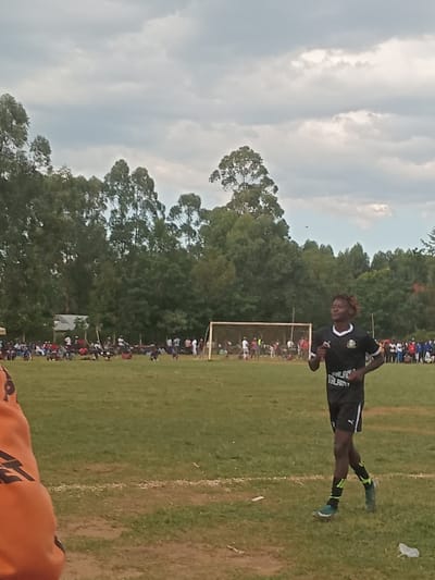 The image depicts an outdoor soccer field in Chevaywa ward, Kenya, under a cloudy sky. In the midground, a male individual with dark skin and braided hair, wearing a black and white soccer uniform (jersey with a green emblem and "MALALA" text, black shorts, black and yellow/green striped socks, and light-colored athletic shoes), runs from right to left across a grassy field. A white line marks the field in the lower-midground. In the background, a white soccer goal is visible. Along the far side of the field, a crowd of spectators is gathered, and behind them, various structures and a dense line of tall green trees extend across the horizon. A partial view of an orange garment is present in the immediate lower-left foreground.