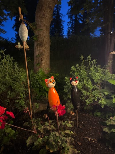 The image depicts a nighttime garden scene in La Villa, Italy. The foreground is dominated by dark soil and lush green foliage, interspersed with vibrant red flowers and buds. Two decorative garden ornaments shaped like cats stand prominently. The cat on the left is orange with a black nose and white detailing on its face, while the cat on the right is black with a red nose and white detailing. Behind the ornaments, a large tree trunk is visible, with its bark textured and a wooden stick extending vertically upwards from the ground, adorned with a pale, elongated ornament at its top. The background shows dense green bushes and trees, with the dark blue of the evening sky visible through the branches. The overall lighting suggests it is dusk or early night, with artificial light likely illuminating the scene.
