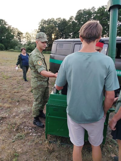 In the city of Олтушскі сельскі Савет, Belarus, a man in camouflage military uniform stands to the right of a green metal structure, possibly a food service unit, with a silver mug on top. He is wearing a camouflage cap and black boots. To his left, a woman in a dark jacket and blue patterned shirt walks away from the camera, holding a white object. In the background, a dark-colored vehicle with a green stripe is visible, and behind it, a line of trees borders a grassy field under a pale sky. Two young men, one with his back to the camera wearing a light blue t-shirt and light shorts, and another partially visible with a red and black hat, are positioned near the green structure and the vehicle. The ground is covered in dry grass and scattered leaves.