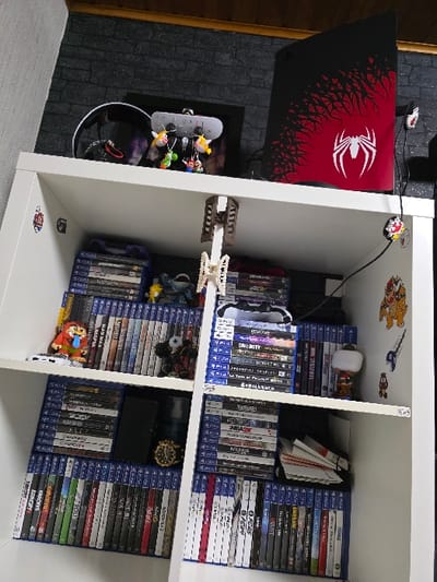 The scene depicts a multi-tiered white shelving unit situated indoors against a dark grey brick-patterned wallpaper, beneath a wooden ceiling trim. The location is an unidentifiable room within the city of Zweibrücken, Germany.

On the uppermost shelf, a black and red PlayStation 5 console, customized with a white Spider-Man logo and red web-like graphics, stands vertically on the right. To its left, a white accessory stand or charging station holds multiple small character keychains, including representations of Yoshi and Mario. A black gaming headset is positioned behind this stand, and a power strip is mounted on the underside of the shelf above it. A black cable with a red and black circular clip descends from the shelf's right edge.

The second shelf displays numerous horizontally stacked PlayStation 4 game cases. A red and orange character figurine with an extended tongue is placed on the left stack, alongside a dark, multi-eyed monster-like figurine. On the right side of this shelf, a white PlayStation controller rests on game cases, and a small Pop! Vinyl-style figurine is present. The vertical wall segment to the right of this shelf features several decals, including a Bowser character and other smaller graphics.

The third shelf also contains horizontally stacked PlayStation 4 game cases. A black smartphone lies flat on the cases to the left, next to a gold-colored wristwatch and a blue glass bottle, possibly containing fragrance. The right section of this shelf holds additional game cases and a stack of white papers or booklets.

The lowest visible shelf is densely packed with PlayStation 4 game cases arranged vertically, showcasing their blue spines across the entire width of the unit. No individuals are present in the image.