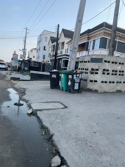The image displays an urban street scene in Ikota, Nigeria, under clear daylight conditions. The foreground presents a broken concrete road surface with a significant, elongated puddle of dark liquid containing debris, including a dark plastic bag and other refuse, on the left. A raised concrete section, possibly a drain cover or part of a damaged sidewalk, is visible adjacent to the puddle.
Towards the center, three wheeled refuse bins stand on the concrete roadside: two black and one teal-green. The black bin on the far left has the word "LOAN" in red. The black bin on the far right has "ICAN FAMILY" and other text visible. All bins appear partially full or overflowing. Immediately behind these bins and extending to the right is a low, weathered concrete block wall featuring multiple rectangular openings arranged in rows. This wall has various posters and graffiti affixed to its surface.
In the midground, a series of multi-story buildings line the street. These structures exhibit modern architecture with varying facades, including white stucco, dark grey panels, and patterned brick/tile accents, typically featuring multiple windows and balconies with dark railings, some equipped with electric or barbed wire security features.
On the left side of the street, further down, utility poles heavily laden with numerous overhead wires are visible, along with multiple commercial signboards. One sign clearly displays "bet9ja," and another "ACCESS." A small structure, possibly a kiosk, stands near these signs. A single individual, indistinctly visible, stands near this kiosk. A white SUV is parked or moving on the road in the distance on the left. A prominent, light-colored utility pole stands vertically in the right foreground, partially obscuring the view of the buildings beyond. The sky above is light blue and clear.