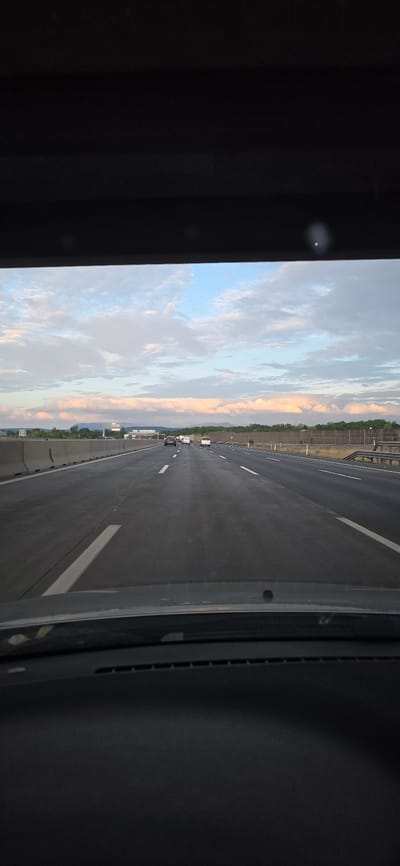 A forward-facing, eye-level shot from the interior of a vehicle looking out onto a highway. The scene captures an overcast sky with clouds tinged pink and white, suggesting either sunrise or sunset. The highway stretches ahead, with multiple lanes and vehicles visible in the distance. The road is dark asphalt, marked with white lines. Guardrails and sound barriers line the sides of the highway, and trees and distant hills form the horizon. The image is taken from the driver's perspective, looking over the dashboard. Based on the provided context of Bad Vöslau, Austria, this scene is likely a section of the Süd Autobahn (A2) or a similar major roadway in the region, possibly near the Austrian Alps. No people are clearly visible.