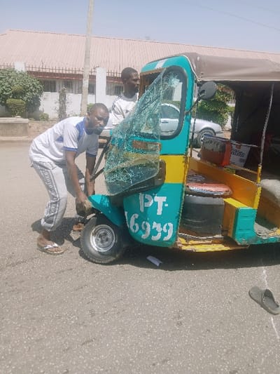 The scene depicts an outdoor daytime incident involving a damaged three-wheeled motorized vehicle on an asphalt road. Two adult males are present in the immediate vicinity of the vehicle.
The vehicle is a teal-green and yellow auto-rickshaw (tuk-tuk) with significant damage to its front. The windshield is shattered into numerous fragments, remaining partially attached to its frame. The front fender appears bent, and some components are dislodged or missing. White text "PT- 6939" is visible on the teal-green bodywork below the windshield. Inside the passenger compartment, various items are stacked, including a box with "Delta" branding.
One male, wearing a white t-shirt with blue trim and grey track pants with white lettering, is crouched low on the left side of the vehicle, near the front wheel. His hands are positioned near the damaged front fender and wheel assembly, suggesting an attempt to inspect or manipulate it. He wears brown flip-flops. The second male, wearing a white long-sleeved shirt with black stripes on the sleeves, stands upright behind the first male, partially obscured by the vehicle's frame and the shattered windshield. His gaze is directed forward.
On the asphalt ground, to the right of the vehicle, a single dark-colored slide sandal lies abandoned. Small pieces of white paper or debris are also scattered near the vehicle's right side. The background features a light-colored building with a dark roof and a perimeter fence topped with barbed wire. Lush green foliage is visible near the fence. A portion of another light-colored vehicle is discernible behind the three-wheeler. The sky is clear and bright, indicating daylight conditions. The specific city of Kwekwe, Zimbabwe, cannot be confirmed from the visual information provided in the image.