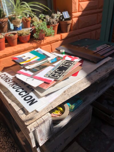 This image captures a sunlit outdoor or semi-outdoor scene, likely a patio or garden area, in Beccar, Argentina. On the left, a terracotta-colored brick wall or facade is visible, supporting several terracotta pots containing a variety of succulents and small plants. One pot features a small solar light, and a garden gnome is discernible among the foliage in another.
Strong, defined shadows indicate bright, clear sunlight, suggesting it is likely midday or early afternoon.
On the right, a rustic wooden pallet or shelf holds a casual arrangement of stacked objects. Prominently displayed is an Argentine license plate, which is upside down but clearly legible, showing the alphanumeric code "GEG 761" and the country name "ARGENTINA". Beneath the license plate, a stack of items includes several framed pictures or small canvases, one of which depicts two people. Additionally, there are white signs with black text; one sign is partially visible but clearly shows "CONSTRUCCION", likely part of a phrase such as "OBRA EN CONSTRUCCION" (Work in Construction). Other miscellaneous items, including a small green spool and a light-colored, round object, are also present within the wooden structure. The scene portrays a casually decorated, lived-in space.