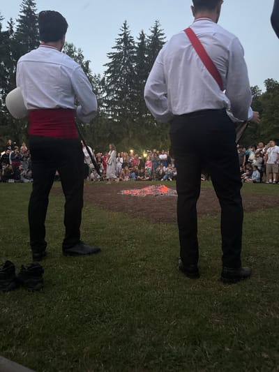 The image shows a group of people gathered outdoors, with a bonfire in the background. In the foreground, two individuals are prominently featured, facing away from the camera. They are dressed in white long-sleeved shirts with red sashes and black pants. The person on the left is holding a white object, possibly a drum or a container, and is wearing dark shoes. The person on the right is also wearing dark shoes.
The crowd in the background is diverse, consisting of men, women, and children, many of whom are watching the bonfire. The setting appears to be a grassy area, possibly a park or a field, with trees visible in the background. The lighting suggests it is late afternoon or early evening. The overall atmosphere seems to be one of a community gathering or a cultural event. No visible text is present in the image. The context of an unknown location in Bulgaria is provided.