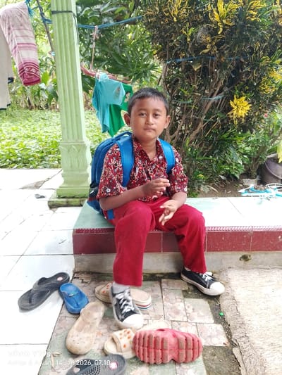 A young boy wearing a red shirt, red pants, a blue backpack, and black sneakers is sitting on a tiled step. Several pairs of sandals and flip-flops are scattered on the ground near his feet. Behind him, there is a green pillar and a lush green bush with yellow flowers. Laundry hangs on a line in the background. The timestamp "VIVO Y19s Artur & Alyan 14 JAN 2026, 07:10" is visible in the lower left corner.