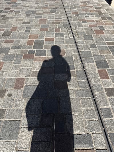 The image shows a shadow of a person cast on a cobblestone pathway in Groningen, Netherlands. The pathway is made of rectangular granite stones of various shades of grey and some reddish-brown stones. A metal drainage grate runs along the right side of the pathway, with some cigarette butts visible within it. The lighting suggests it is daytime, and the shadow indicates the person is standing and facing away from the sun.