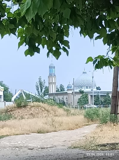 The image shows a view of a mosque in Tokmok City, Kyrgyzstan, through overhanging green foliage. The mosque features a large, patterned dome and a tall, slender minaret with blue accents. In front of the mosque, there is a white, arched structure that appears to be part of a playground or a decorative element. The foreground is a dusty, unpaved path leading towards the mosque, with dry, overgrown grass on either side. The sky is a pale blue. A date and time stamp in the bottom right corner reads "22.06.2026 13:01".