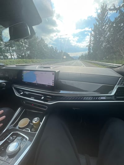 The image depicts the interior of a modern BMW vehicle, viewed from the front passenger seat, while traveling on a multi-lane highway in the Municipal Unit of Pelasgia, Phthiotis, Greece. The vehicle's digital dashboard displays a speed of "122" (likely km/h) on the left and a navigation map centered on the Maliakos Gulf coastline, indicating nearby locations such as Atalanti and Thermopylae. The dashboard features carbon fiber trim and an illuminated "///M" logo on the passenger side. In the center console, two cupholders are visible, containing a beige paper cup and a silver/yellow beverage can. The lower legs of the passenger, dressed in dark trousers, are visible on the left, along with a partial view of their hand. The lower legs of the driver, also in dark trousers, are partially visible on the far right. Outside the vehicle, the highway is bordered by dense coniferous trees under a partly cloudy sky with bright sunlight causing significant glare on the windshield. Streetlights line the road, and distant mountains are visible on the horizon.