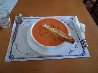A bowl of tomato soup is presented on a white plate, topped with a piece of toasted bread drizzled with olive oil. The bowl rests on a white placemat with blue decorative borders and text. The placemat is adorned with the phrases "Bon Profit" and "Bom Apetite" on opposite sides, and "Enjoy your Meal" in the lower left corner. To the left of the bowl is a metal spoon, and to the right, a folded white napkin with cutlery tucked inside. A clear glass of water is visible to the far left. The entire setting is placed on a wooden table. The image appears to be taken indoors, likely in a restaurant in Mas Aliu, Spain, given the Spanish phrases on the placemat.