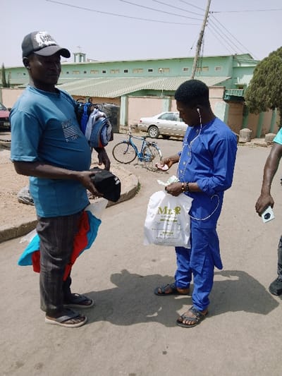 The scene depicts an outdoor transaction occurring in the city of Mumbwa, Zambia. Two adult males are centrally positioned, engaged in an exchange. The male on the left, wearing a dark baseball cap, light blue t-shirt, dark plaid trousers, and flip-flops, holds various folded garments (blue, white, red, and a black item) suggesting he is a vendor. The male on the right, wearing a royal blue tunic, matching trousers, and dark sandals, is holding small denomination banknotes (pink and green) in his right hand and a white plastic "Mr. R" shopping bag in his left hand. He is also wearing white in-ear headphones. A third male's lower right arm and hand, holding a white smartphone, are partially visible on the far right.
In the background, a light-colored sedan is parked on the street, behind which is a light green building with a corrugated roof and a visible white cross, indicating a religious institution. A light pink wall encloses the property. A blue bicycle with a front basket is parked near a curb between the two primary figures and the background buildings. A wooden utility pole with multiple wires stands on the right side of the frame. The ground is paved, and long shadows are cast, indicative of daylight conditions.