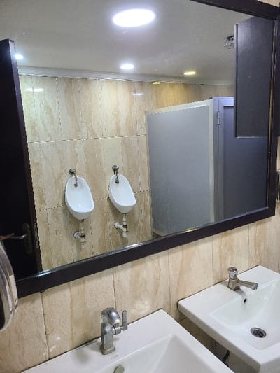 The image displays the interior of a public restroom located in Ibeju, Nigeria. The primary view is from the perspective of someone facing a large, dark-framed rectangular mirror. In the immediate foreground, two white ceramic vanity sinks are visible, each fitted with a silver-toned single-lever faucet. The wall behind the sinks and visible in the reflection is clad with light-toned, mottled ceramic tiles. Reflected in the mirror are two white ceramic wall-mounted urinals, each equipped with exposed silver-toned plumbing. Beyond the urinals, light grey, opaque privacy partitions for toilet cubicles are reflected, alongside a closed, dark-colored cubicle door. The ceiling, also seen in the reflection, contains multiple recessed circular light fixtures emitting bright white illumination. A partial view of a dark-colored door handle and a silver-toned wall-mounted dispenser are visible on the far left side of the frame, outside the main mirror reflection. No individuals are present in the scene, and no active interactions are depicted.