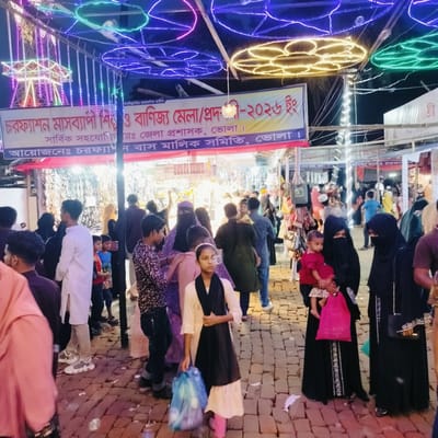 The image captures a bustling night scene at the "Charfasson Month-long Industrial and Trade Fair/Exhibition" in Charfasson, Bhola, Bangladesh.

**Subjects:** A large crowd of people, including men, women, and children, are walking through the fair. Many women are dressed in traditional attire, with some wearing hijabs or niqabs. Children are seen with adults; one child in a red top is held by a woman in a black abaya, and another child is on an adult's shoulders. A young girl in a cream top, black scarf, and pink trousers holds a blue plastic bag while looking towards the viewer. Most faces are either turned away or obscured by the crowd.

**Objects and Setting:** The fair is illuminated by an abundance of artificial lights. Overhead, colorful circular string lights in blue, green, and yellow create a festive atmosphere. In the background to the left, parts of a brightly lit Ferris wheel are visible. The ground is paved with reddish-brown cobblestones. Market stalls line both sides of the pathway, displaying various merchandise, though specific items are not clearly discernible beyond bags and small decorative pieces.

**Activity or Event Taking Place:** People are engaged in strolling through the fair, observing the stalls, and likely shopping or socializing. The scene depicts a lively public gathering during an evening event.

**Notable Details:** The time of day is night, emphasized by the dark sky and the reliance on artificial lighting. The overall mood appears busy and festive. The location is clearly identifiable from a large banner stretched across the upper part of the scene.

**Visible Text:** A prominent banner in Bengali provides details about the event:
*   "বিস্মিল্লাহির রাহমানির রাহীম" (In the name of Allah, the Most Gracious, the Most Merciful)
*   "চরফ্যাশন মাসব্যাপী শিল্প ও বাণিজ্য মেলা/প্রদশী-২০২৬ ইং" (Charfasson Month-long Industrial and Trade Fair/Exhibition - 2026 AD)
*   "সার্বিক সহযোগি য়েঃ জেলা প্রশাসক, ভোলা।" (Overall cooperation/assistance: District Administrator, Bhola.)
*   "আয়োজনেঃ চরফ্যাশন বাস মালিক সমিতি, ভোলা।" (Organized by: Charfasson Bus Owners' Association, Bhola.)