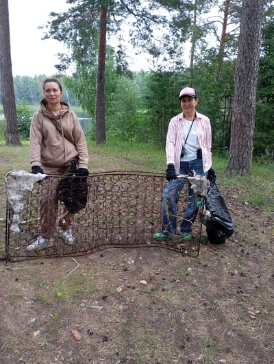 Two women are standing in a wooded area, next to a large, rusty bed spring frame. The woman on the left is wearing a tan hoodie and brown pants. She has black gloves on and is holding a black trash bag. The woman on the right is wearing a pink and white striped shirt, a white t-shirt underneath, blue jeans, and a pink baseball cap. She also has black gloves on and is holding a black trash bag. Both women are looking at the camera and appear to be engaged in a cleanup effort, as evidenced by the trash bags and the discarded bed frame. The background shows a lake and a dense forest with tall trees. The ground is covered in pine needles and dry grass. The lighting suggests it is daytime, likely overcast. The overall mood seems neutral. There is no visible text in the image. This scene likely takes place in Vekoshinka, Russia, given the provided context.