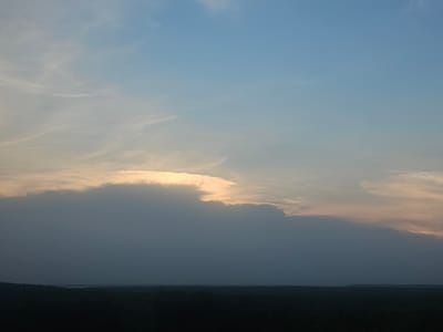 A wide shot captures a dusky sky above a silhouetted landscape, likely the city of Taezhnoye, Russia. The sky is dominated by a gradient of soft blues and pale oranges, with wispy clouds streaking across the upper half. A thick, dark cloud formation stretches across the lower half of the sky, obscuring the horizon. The sun appears to be setting or rising behind this cloud, casting a bright, golden glow on its upper edge. Below the sky, a dark, indistinct line represents the tree line of the Taezhnoye landscape, with a sliver of lighter sky or water visible on the far horizon. The overall impression is one of a serene, natural landscape at twilight.