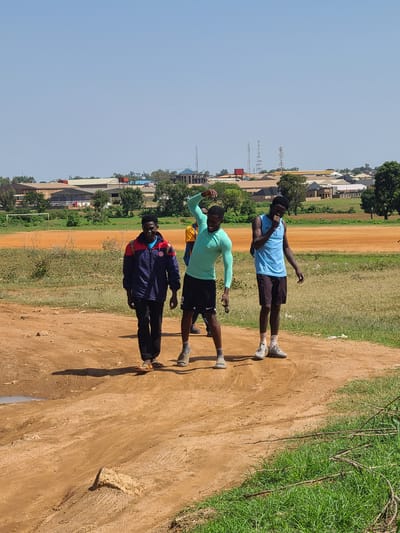 In Barikin Jirga Summa, Nigeria, three young men are captured mid-stride on a dusty, dirt path. The man on the left, wearing a blue, red, and white jacket and black trousers, smiles at the camera. The central figure, clad in a teal long-sleeved athletic shirt and black shorts, strikes a pose with his arm raised. To the right, a man in a blue tank top and dark shorts, with a backpack, appears to be eating something. The background reveals a rural or semi-urban landscape with a large, orange, dusty field, possibly a sports field, bordered by green grass and trees. Beyond that, buildings and communication towers are visible under a clear blue sky.