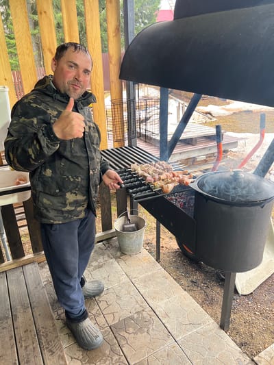 A man stands on a wooden deck beside a black, barrel-shaped barbecue grill. He wears a camouflage jacket, dark pants, and gray boots. He gestures with a thumbs-up. Skewers of meat are cooking on the grill. The environment includes wooden structures and ground covered with paving stones and vegetation. The location may be Bol'shakino, Russia.
