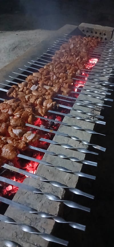 In Daliguzar, Uzbekistan, skewers laden with seasoned meat are being grilled over glowing red coals. The meat, cut into uniform pieces, is arranged densely on long metal skewers. The skewers are supported by a structure made of bricks, positioned over an outdoor fire pit. Smoke rises from the coals and the grilling meat, indicating the active cooking process. The scene is illuminated by the fire's glow, suggesting it is nighttime or a dimly lit area. The background is dark and indistinct, with some sandy or dusty ground visible. The overall impression is of a traditional outdoor barbecue.